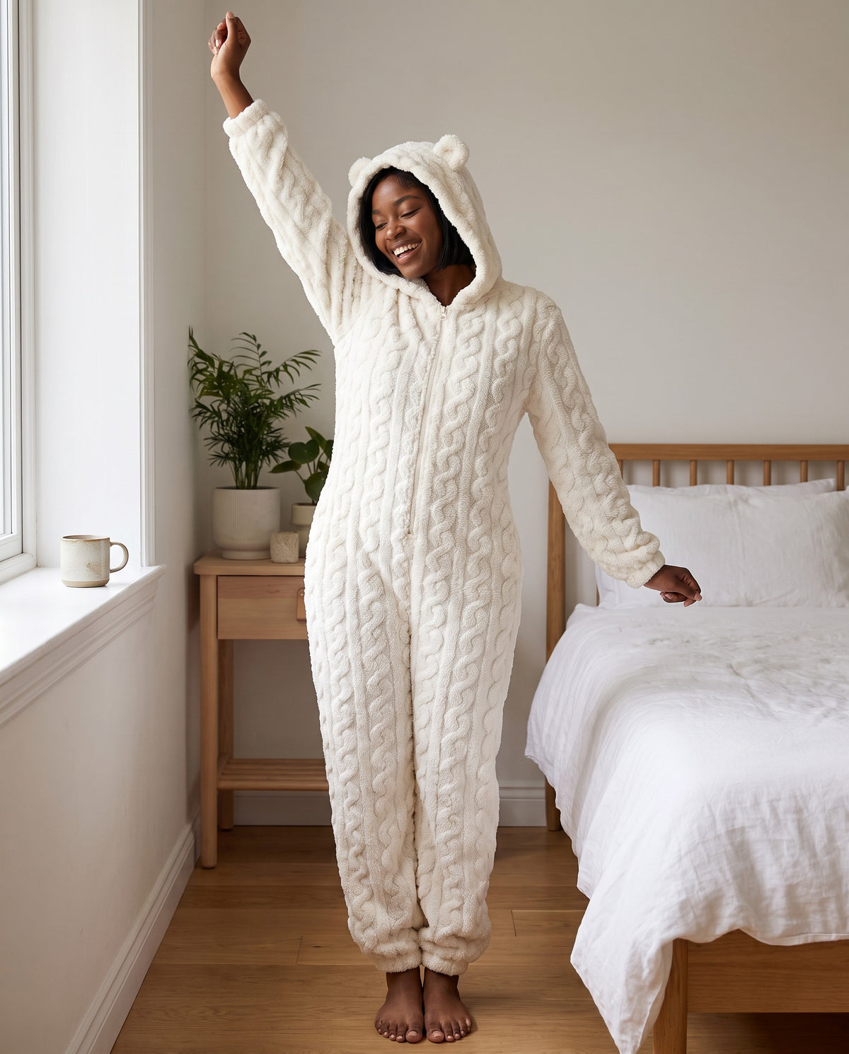 Woman stretching in a bright bedroom in the TEDDY Cozy Bear Onesie in White, hood up with bear ears